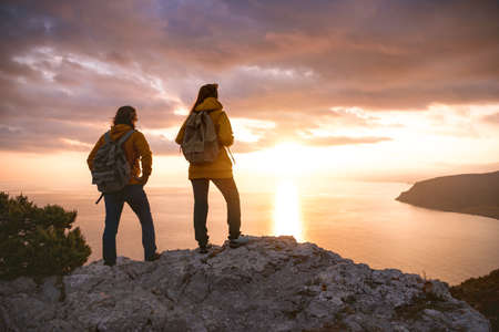 Two hikers with backpacks looks at sea sunset from view pointの写真素材