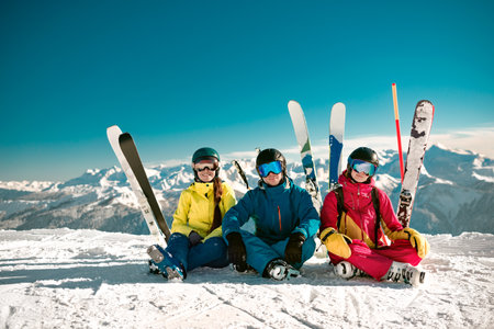 Three happy young skiers sits at mountain top and smiles. Winter vacations concept at ski resortの写真素材