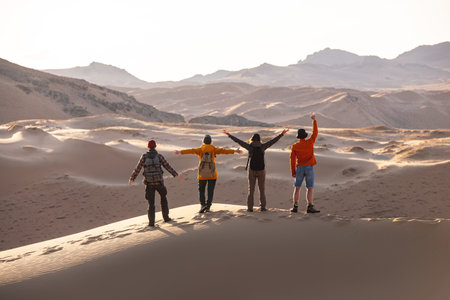 Four happy tourists with backpacks stands on sandy dune with open arms and looks at sunsetの写真素材