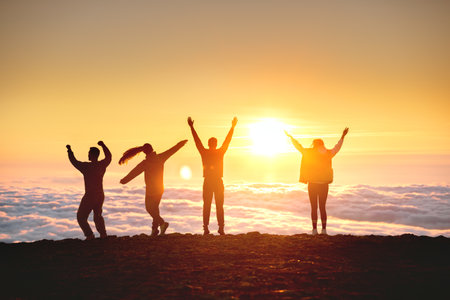 Group of happy tourists are having fun and dance at sunrise on mountain top over cloudsの写真素材