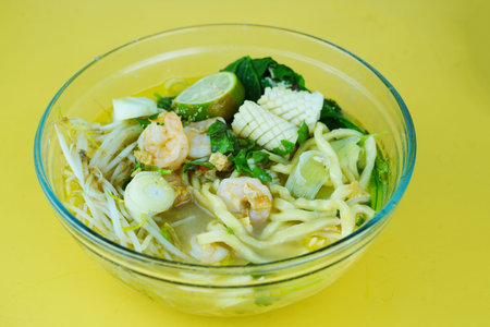 A clear glass bowl of seafood noodle soup with shrimp, squid, and vegetables. Food Photography