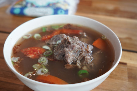 A bowl of ribs soup with meat, tomatoes, carrots, and scallions served on a wooden table.の写真素材