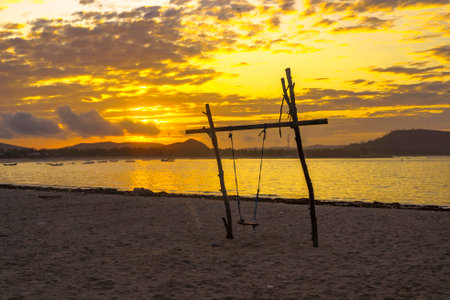 A breathtaking sunrise paints the sky above a secluded beach near Mandalika, Lombok, Indonesia casting a golden glow on the calm waters and highlighting a lone swing on the shore.の写真素材
