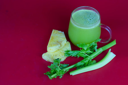 a bright green colored celery and pineapple juice on a red table. There is a sliced piece of...