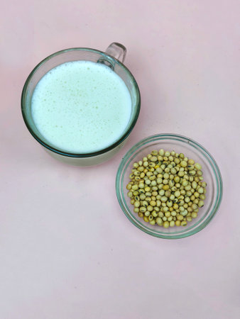 A glass of frothy soy milk beside a bowl of yellow soybeans on a pink background.の写真素材