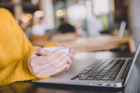 Woman in yellow patterned, woolen sweater sitting with laptop and  holding a mug of coffee on the...