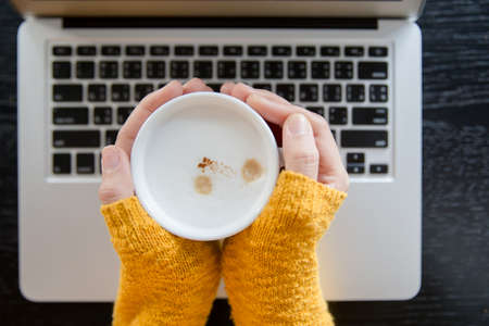 Woman in yellow patterned, woolen sweater sitting with laptop and  holding a mug of coffee on the...