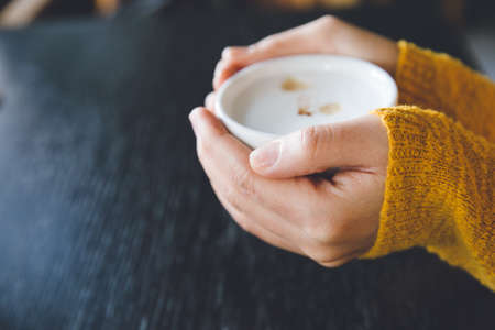 Woman holding cup of hot coffee on dark wooden table, closeup photo of hands in warm sweater with...