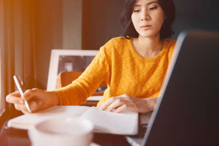 Young asian business woman in yellow dress sitting at table in cafe and writing in notebook. On table is laptop, smartphone and cup of coffee. Freelancer working in coffee shop. Student learning online.の写真素材
