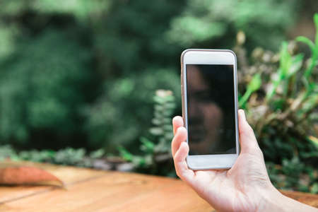Female hand holding a moblie phone device on a wood table with green natural morning background.の写真素材