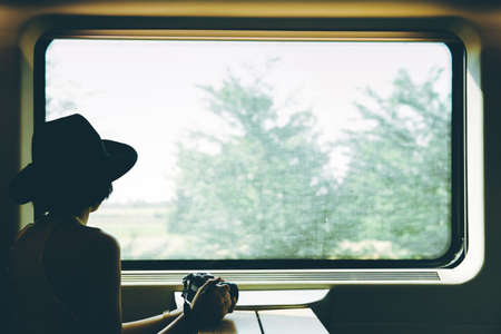Enjoying travel. Young pretty woman traveling alone by train, holding a camera and looking at the view through the window.の写真素材