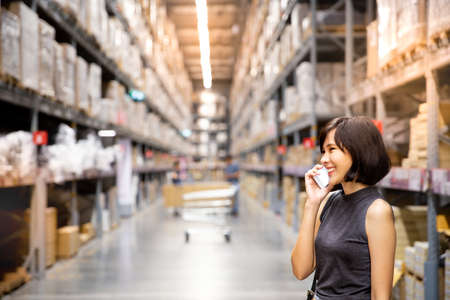 An asian businesswoman having a phone conversation in the warehouse. Cart and boxes on rows of shelves in warm light warehouse background.  shopping concept.の写真素材