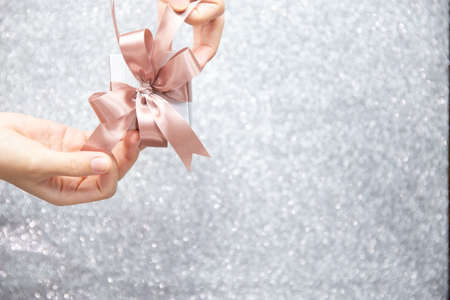 Female hands holding or pulling ribbons of a small gift box with copy space for text and silver glitter background.の写真素材