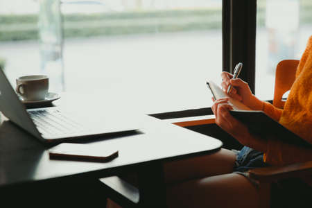 Young business woman in yellow dress sitting at table in cafe and writing in notebook. On table is laptop, smartphone and cup of coffee. Freelancer working in coffee shop. Student learning online.の写真素材