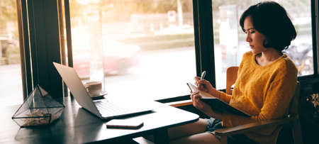 A hipster asian woman working with laptop and writing information into notebookの写真素材