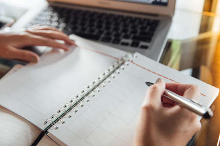 Young business woman in casual dress sitting at table in cafe and writing in calendar notebook. On table has laptop, notebook. Freelancer working in coffee shop. Student learning online.の写真素材