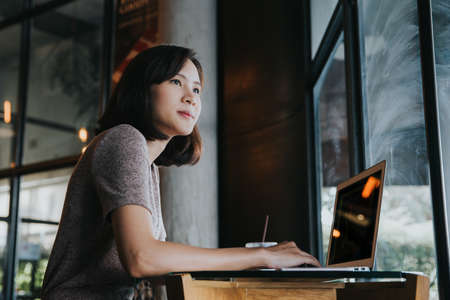 Beautiful young Asian woman working at a coffee shop with a laptop. Female freelancer connecting to internet via computer. Casual Thinking Browsing Concept.の写真素材
