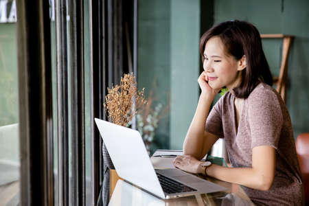 Beautiful young Asian woman working at a coffee shop with a laptop. Female freelancer connecting to internet via computer. Casual Thinking Browsing Concept.の写真素材