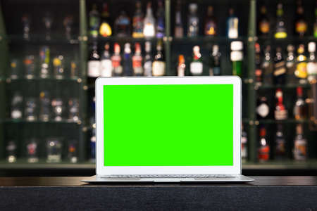 Blank screen of Laptop on blur alcohol drink bottle on bar counter in the dark night background. for create Background or presentationの写真素材
