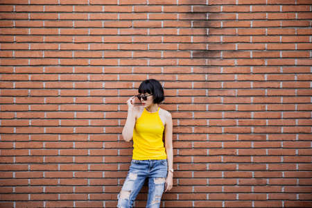 Fashion happy smiling hipster cool girl in sunglasses. Summer posing at urban brick wall background in yellow shirt, jeans and sunglasses. Copy space for text and product presentation.の写真素材