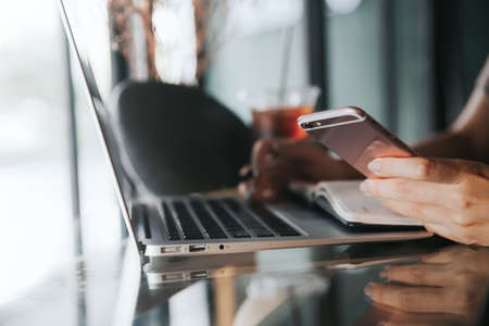 Close up of female hands using smart phone while working on computer laptop. Freelancer typing text...