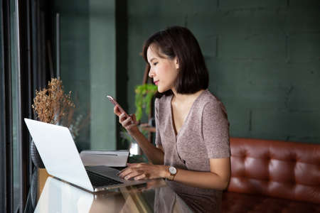 Young beautiful asian woman working in loft cafe or office. Holding smart phone and using laptop computer for browsing and searching internet wifi. Business woman or freelancer working on new project.の写真素材