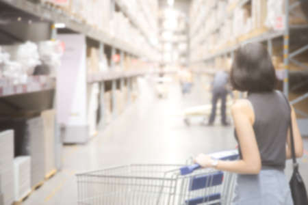 Blurred image of An asian woman doing shopping  and walking with her cart in cargo or warehouse. Boxes on rows of shelves in warm light warehouse background.  Using for Mock up template for craft display of your design,Banner for advertise of product.の写真素材