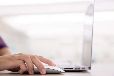 Close up of young female hands using mouse remote for laptop in modern office near by the window. Working on new project with notebook computer flare light, blurred office building backgroundの写真素材