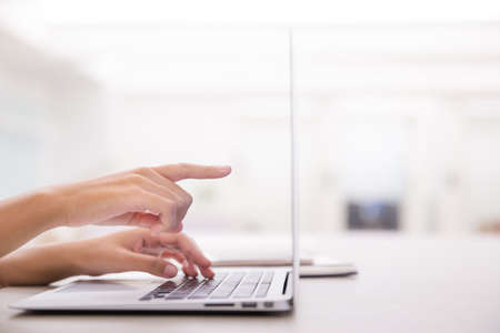 Close up of young female hands using laptop and pointing the screen in modern office or loft near by the window. Working on new project with notebook computer flare light, blurred office building backgroundの写真素材