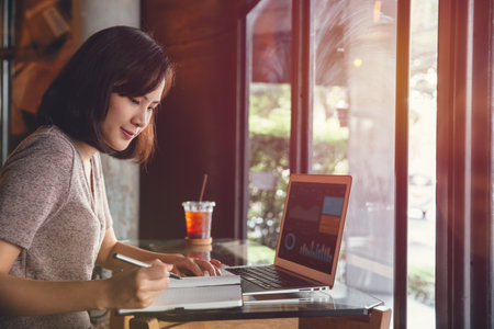Isolated mockup image of laptop and Young business woman in casual dress sitting at table in cafe and writing in notebook. Freelancer working in coffee shop. Student learning online.の写真素材