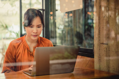 Asian woman using laptop in coffee shop cafe, warm colour. Freelancer working at a cafe with a laptopの写真素材
