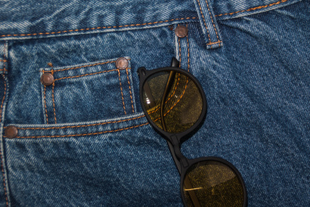 Round sunglasses on a denim texture background. round yellow glasses in the front pocket of jeans. Rough denim fabric. Classic jeans five pockets close-up. Fashionable round glasses in your pocket. Fashion casual style.の写真素材