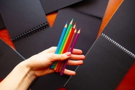 A hand holding bright colored pencils in front of black paper sketch boooks.