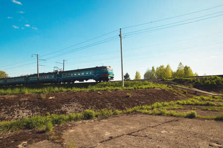 Pleasant sunny summer landscape: old blue sky and clouds, green grass and trees, railway path road, old rusty industrial metal construction and a train. A bright scenery view and spirit of adventure.の写真素材