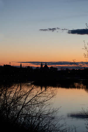 Sunset on Dnieper river. Beautiful Orange cloudy sky, city and church silhouettes reflecting in the river. Pleasant vertical spring photo taken in Rechytsa, Belarus.の写真素材