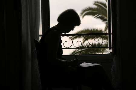 Mature woman writing in her diary in front of the window just after sunset.の写真素材