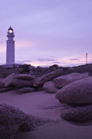 Several stones at the beach with a purple sunset plus a blurred lighthouse in the distanceの写真素材