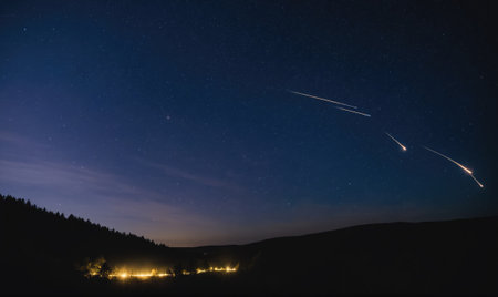 A series of shooting stars streak across the night sky above a dark, wooded hillsideの素材
