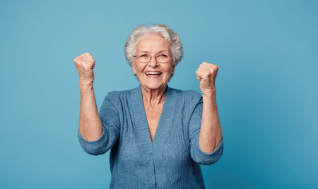 A woman in a blue sweater smiles and raises her fists in celebration against a light blue backgroundの素材