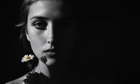 A woman with dark hair holds a single daisy in front of her face, bathed in light against a dark backgroundの素材