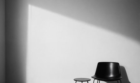 A black chair and a small table sit in a white room with a single, diagonal beam of sunlightの素材