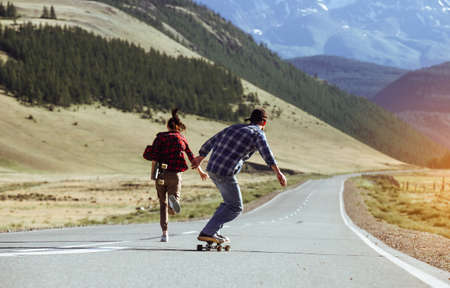 Happy young couple having fun with longboard on the road. Young man and woman skating together on a sunny day in mountain.の写真素材