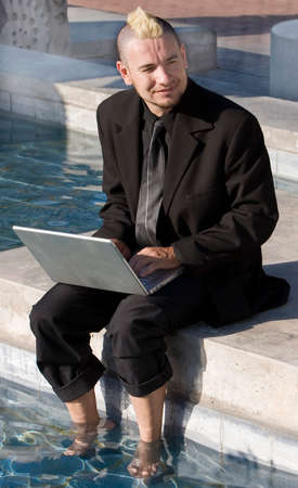 Businessman with a punk haircut works on a laptop computer with his feet in a fountain.の写真素材