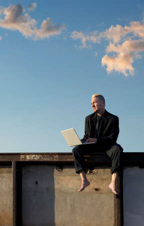 Barefoot businessman with a punk haircut works on his laptop computer sitting on a wall.の写真素材