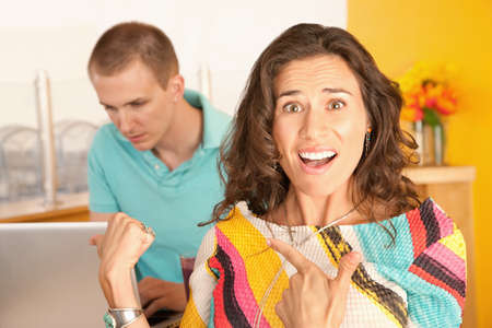 Image of a woman in the foreground with a frustrated expression is pointing to a man in the...