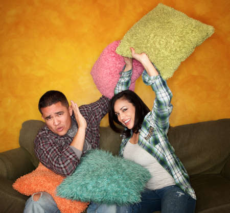 Hispanic couple on couch play fighting with pillows