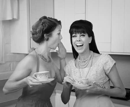 Caucasian woman whispers joke to friend in her kitchenの写真素材
