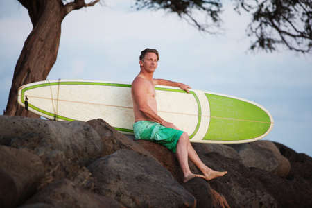 Single adult surfer male outdoors sitting with surfboard on rocksの写真素材