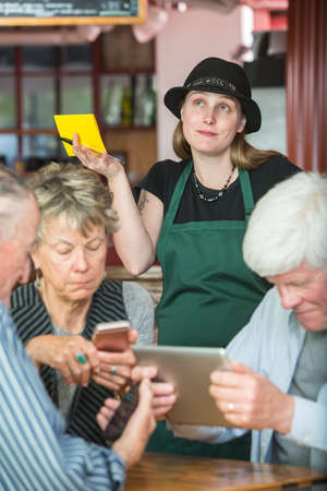 Mature friends in a coffee house distracted by their devices as server waitsの写真素材