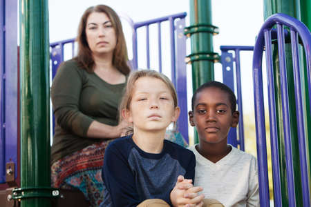 Two male children sitting on playground set with mother behind themの写真素材